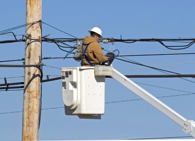 Utility worker in white hard hat and brown jacket repairing power lines from a bucket truck lift against a clear blue sky.
