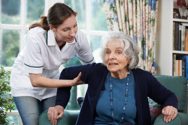 Care Worker Helping Senior Woman To Get Up Out Of Chair