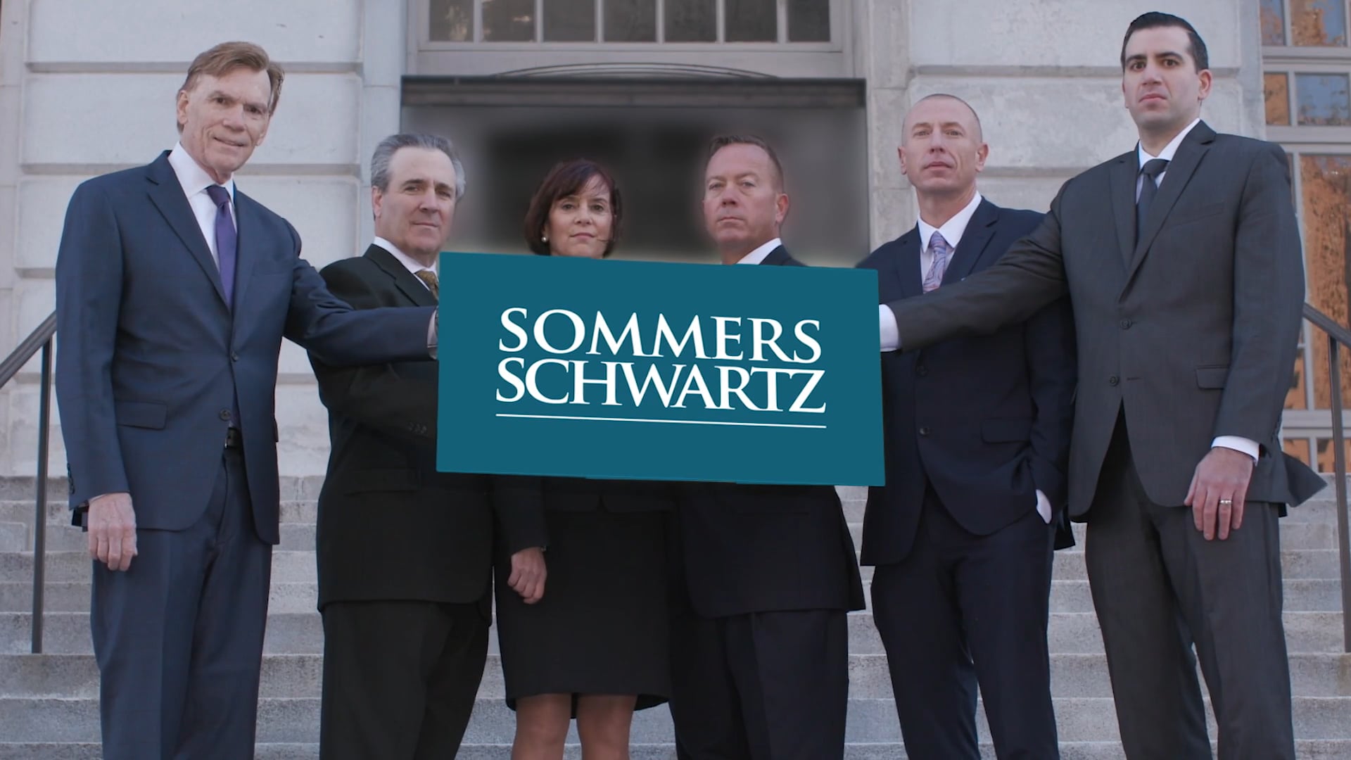 Six attorneys in dark suits standing on courthouse steps holding a teal Sommers Schwartz law firm sign