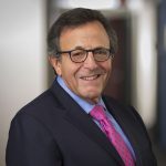 Smiling middle-aged man with dark-rimmed glasses, navy suit, light blue shirt, and pink patterned tie in a professional he...