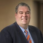 Smiling man in navy pinstripe suit, light blue dress shirt, and orange and blue striped tie against a neutral background
