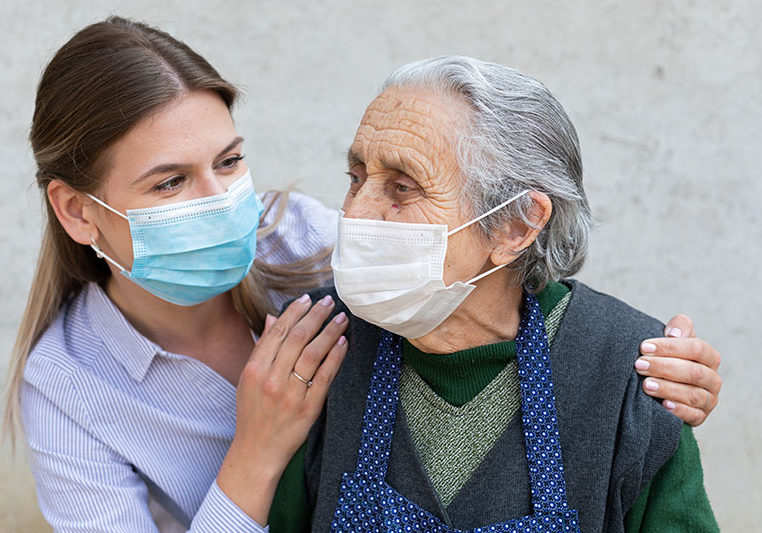Portrait of friendly caregiver posing with elderly ill woman wearing surgical mask because of covid-19 pandemic