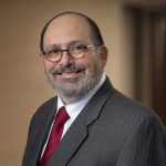 Smiling middle-aged man with glasses and gray beard wearing a gray pinstripe suit and red tie against a neutral background.