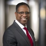 Smiling man in gray suit, white dress shirt, and red tie wearing rectangular glasses, professional headshot in office hallway