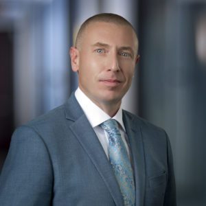 Man in gray suit and blue patterned tie with white dress shirt, professional headshot with blurred office background