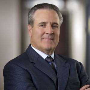 Middle-aged man with gray hair and blue eyes, arms crossed, wearing a dark blue suit and purple striped tie, professional ...