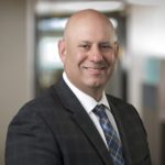 Smiling bald man in a dark gray suit, white dress shirt, and blue plaid tie, posed in a professional office setting.
