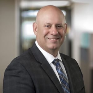Smiling bald man in a dark gray suit, white dress shirt, and blue plaid tie, posed in a professional office setting.