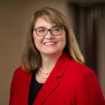 Smiling woman with brown hair and glasses wearing a red blazer, black top, and pearl necklace in a professional headshot p...