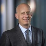 Bald man smiling in a professional headshot, wearing a dark gray suit, light blue shirt, and patterned gray tie against a ...