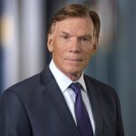 Older man in dark gray suit and blue tie with white dress shirt, professional headshot with blurred office background