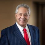 Smiling older man with gray hair wearing a navy blue pinstripe suit, white dress shirt, and red tie against a neutral back...